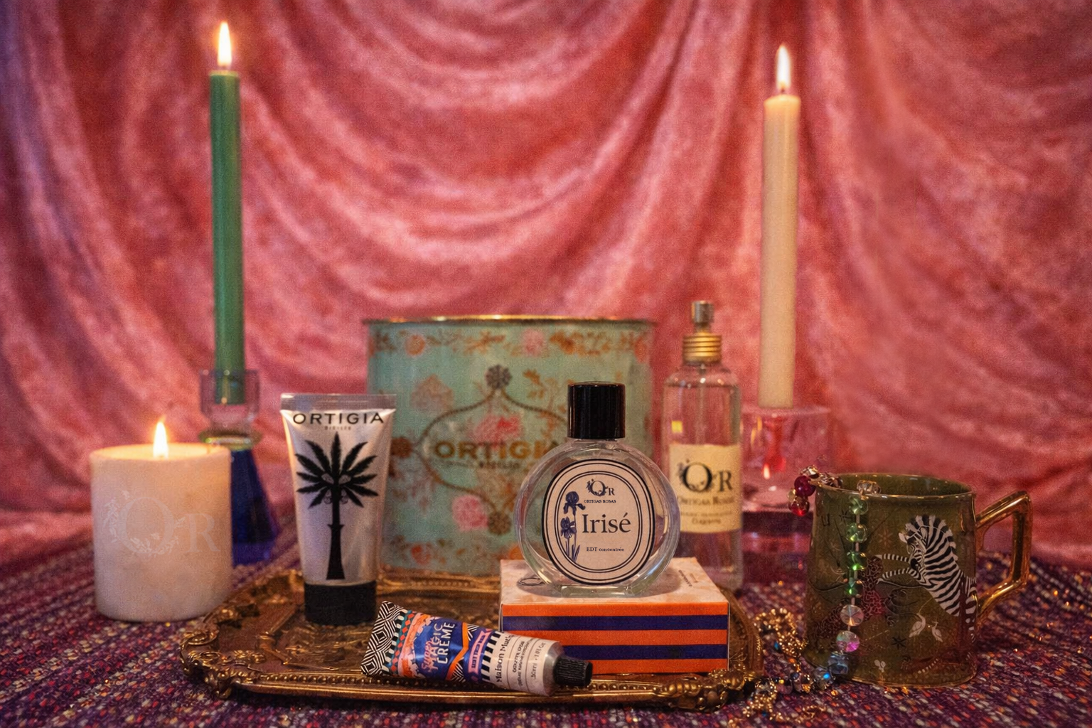 Collection of decorative items including candles, bottles, and a mug on a textured surface with a pinkish-red curtain background, with perfumes and candles for san valentine