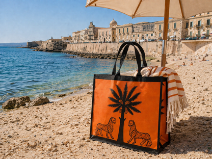 Orange tote bag with palm tree and leopards design on a beach with ocean and buildings in the background