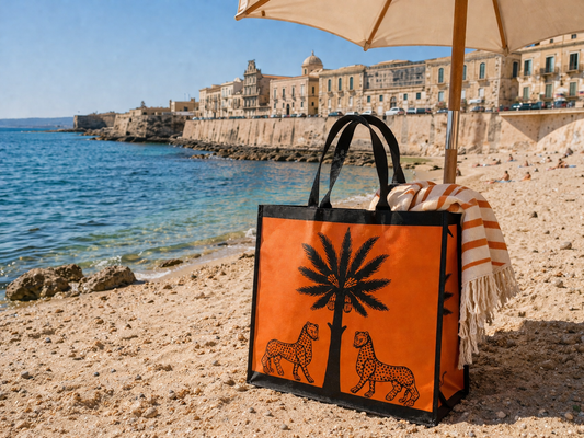 Orange tote bag with palm tree and leopards design on a beach with ocean and buildings in the background